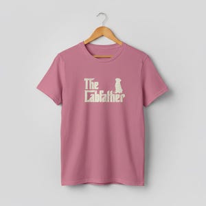 Op de afbeelding: Een stoffig roze t-shirt met de tekst "The Labfather" in een verweerde, gebroken witte lettertype. Een silhouet van een hond staat naast de tekst. Het shirt hangt aan een houten hanger.