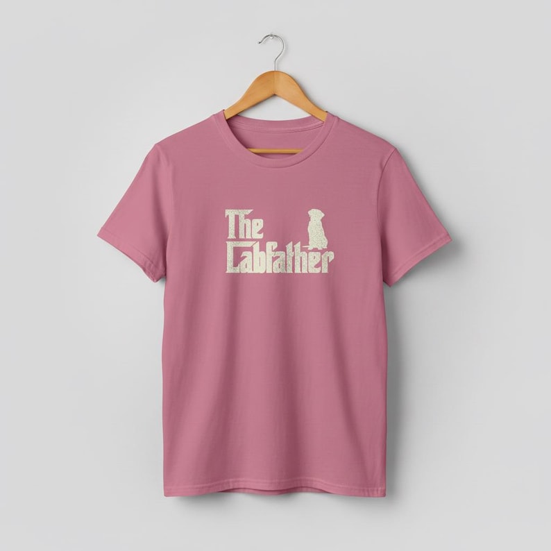 Op de afbeelding: Een stoffig roze t-shirt met de tekst "The Labfather" in een verweerde, gebroken witte lettertype. Een silhouet van een hond staat naast de tekst. Het shirt hangt aan een houten hanger.