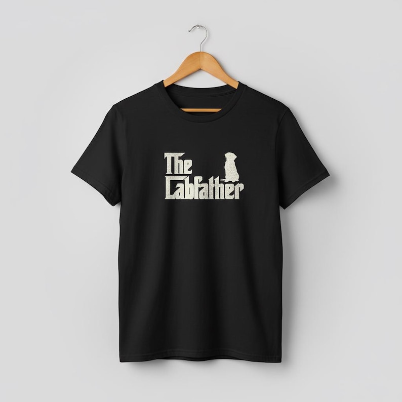 Op de afbeelding: Zwart T-shirt met de tekst "The Labfather" en een silhouet van een hond in vintage stijl. Het shirt hangt aan een houten hanger. Het ontwerp is in een distressed stijl, met een neutrale achtergrond.