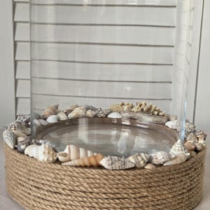Könnte beinhalten: Ein klarer Glas-Windlicht-Halter mit einem Sockel, der mit Naturseil umwickelt ist. Der Sockel ist mit verschiedenen Muscheln und kleinen Steinen verziert. Der Halter ist für eine Kerze oder andere dekorative Gegenstände konzipiert.