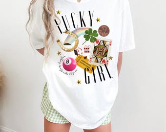May include: White oversized t-shirt with the text "LUCKY GIRL" in black. The shirt features a graphic design of lucky charms, including a horseshoe, a four-leaf clover, a rainbow, dice, a playing card, a penny, and a billiard ball.