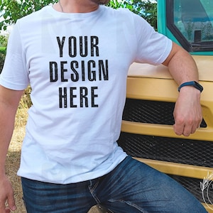 Può includere: T-shirt bianco girocollo con la scritta "YOUR DESIGN HERE" in nero. Il modello indossa jeans blu e un braccialetto blu. Lo sfondo comprende un veicolo giallo e vegetazione.