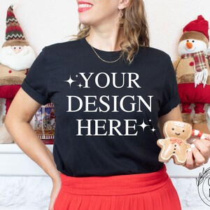 May include: A woman wearing a black t-shirt with the text "YOUR DESIGN HERE" in white. She is holding a gingerbread man cookie in her right hand. The background is decorated with Christmas ornaments.