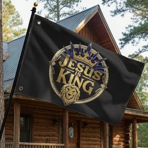 Könnte beinhalten: Schwarze Flagge mit dem goldenen Schriftzug "JESUS IS KING", einer goldenen Krone und einem Löwenkopf-Emblem. Die Flagge wird vor einer Holzhütte gezeigt.