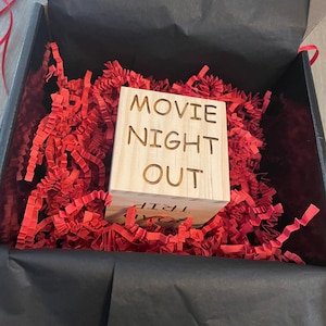 May include: A wooden box with the words "Movie Night Out" carved into the front. The box is sitting in a black box filled with red shredded paper.