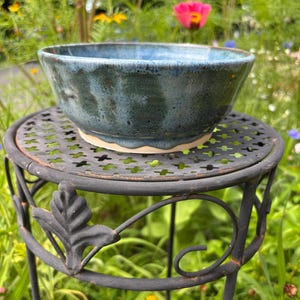 May include: A round ceramic bowl with a glossy glaze featuring a blend of deep blue, teal, and dark green tones. The bowl has a smooth, curved interior and a slightly flared rim, resting on a circular base with a visible light-colored clay edge.