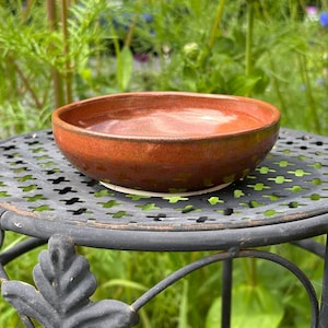 May include: A small, round, ceramic bowl with a glossy, deep orange-red glaze. The bowl features a smooth, slightly uneven rim and a shallow interior basin. The exterior surface shows subtle variations in color, transitioning to a darker tone.