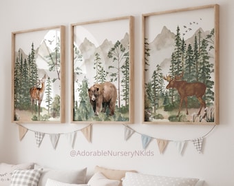 Op de afbeelding: Drie ingelijste wandkunstprints met aquarelillustraties van een hert, een bruine beer en een eland in een boslandschap met bergen. De prints zitten in lichte houten lijsten met een slinger van stoffen vlaggetjes die eronder hangt.
