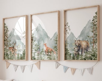 Op de afbeelding: Een set van drie ingelijste wandkunstwerken met aquarelillustraties van bosdieren en bergen. De linker print toont herten, de middelste print een vos en een konijn, en de rechter print een beer met welp in een dennenbos.