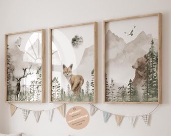 May include: Three framed wall art prints featuring watercolor illustrations of a deer, a fox, and a bear in a forest landscape with mountains and trees. The prints are displayed in light wood frames with a string of fabric bunting hanging below.
