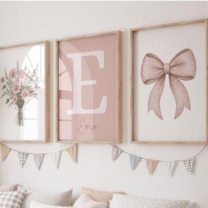 May include: Three framed wall art prints in light wood frames. The left print features a bouquet of pink flowers. The centre print displays a large white letter E and the name Ema in script. The right print shows a pink polka dot bow.