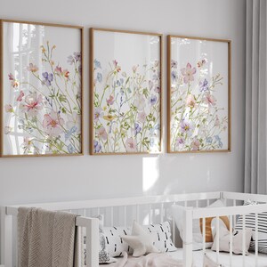 K&ouml;nnte beinhalten: Drei gerahmte Aquarellbilder mit rosa, lila und blauen Blumen auf wei&szlig;em Hintergrund. Die Rahmen sind aus hellem Holz.