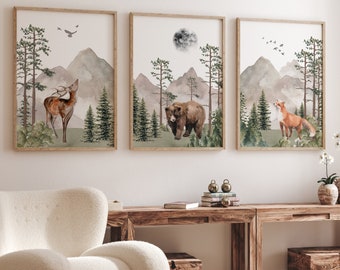 Op de afbeelding: Een set van drie ingelijste wandkunstprints met aquarelillustraties van een hert, een beer en een vos in een boslandschap met bergen, dennenbomen en een maan. Elke rechthoekige print is gevat in een lichtgekleurde houten lijst.