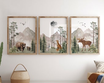 Op de afbeelding: Een set van drie ingelijste wandkunstprints met aquarelillustraties van bosdieren. De linkerprint toont een beer met jong, de middelste een vos en konijn met een maan, en de rechter een eland, allemaal afgebeeld tegen bergen en dennenbomen.