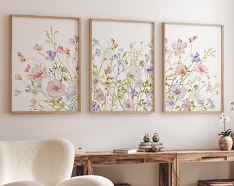 May include: Three framed watercolor paintings of pink, purple, and blue wildflowers on a white background. The paintings are arranged in a row on a white wall.