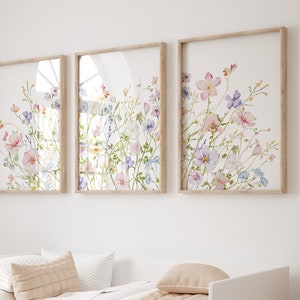 K&ouml;nnte beinhalten: Drei gerahmte Aquarelldrucke von rosa, lila und blauen Wildblumen auf wei&szlig;em Hintergrund. Die Drucke sind in einer Reihe an einer wei&szlig;en Wand angeordnet.