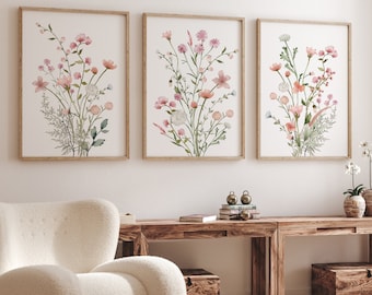 May include: Three framed watercolor paintings of pink and white flowers with green stems and leaves on a white wall. The paintings are in wooden frames and are hung in a row.