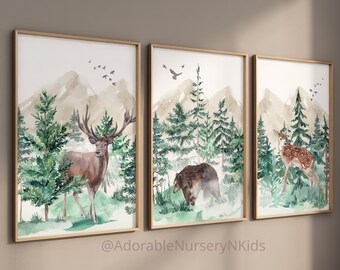 Op de afbeelding: Een set van drie ingelijste aquareldrukken met bosscènes. De eerste toont een hert, de tweede een bruine beer en de derde een reekalf, allemaal tegen een achtergrond van groene dennenbomen, bergen en vogels in de lucht.