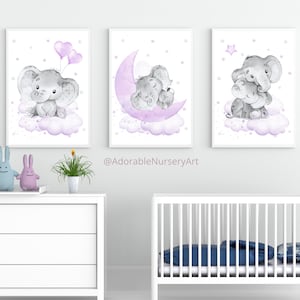 May include: Three watercolour prints featuring grey elephants with purple accents on a white background. The first print shows an elephant holding two purple balloons. The second print shows an elephant and a baby elephant sleeping on a purple crescent moon. The third print shows an elephant holding a baby elephant with a purple star in the background. The prints are displayed on a white wall above a white dresser and a white cot.