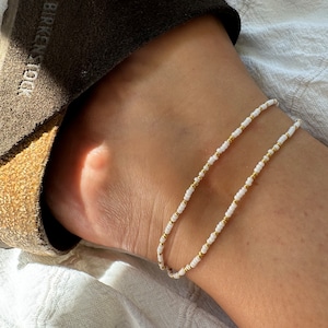 Könnte beinhalten: Ein weißes und goldenes Fußkettchen. Das Armband besteht aus kleinen, runden Perlen, die auf einer dünnen Schnur aufgereiht sind. Die Perlen sind weiß mit goldenen Akzenten.