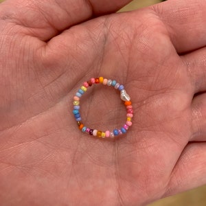 Könnte beinhalten: Ein bunter Perlenring mit einer einzelnen Perle. Der Ring besteht aus kleinen, bunten Perlen in den Farben Rosa, Blau, Gelb, Orange und Rot.