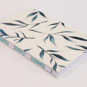 May include: A white notebook with a blue and green watercolor leaf pattern on the cover. The notebook is closed and lying on a white surface.