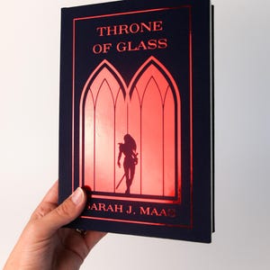 Puede incluir: Un libro de tapa dura titulado "Throne of Glass" de Sarah J. Maas. El libro tiene una cubierta azul marino con letras rojas y una ilustración roja de una figura con una espada. El libro está sostenido en una mano.