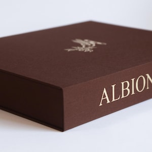 Puede incluir: Una caja rectangular de color marrón con un acabado texturizado. El texto ALBION BELL está impreso en dorado en el lateral. Un pequeño emblema dorado con un pájaro está centrado en la superficie superior. La caja tiene una tapa plana y bordes.