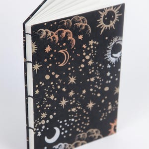 Puede incluir: Un cuaderno negro con un patrón celestial dorado y blanco. La portada presenta estrellas, lunas y soles.
