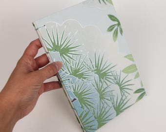 May include: A small, blue and green journal with a floral pattern. The journal is bound with a light blue thread.