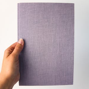 Puede incluir: Un libro rectangular con una cubierta texturizada de color lavanda. El libro está sostenido por una mano sobre un fondo blanco. La cubierta tiene una apariencia de tejido.