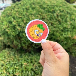 May include: A round sticker with a red border and a white background. The sticker features a cartoon illustration of a plate of food with a yellow and orange color scheme. The food is topped with a green garnish.
