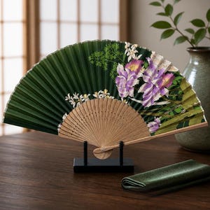 May include: A folding hand fan featuring a deep green background with a printed illustration of purple and white flowers. The fan has a light-colored wooden frame with intricate cut-out details and is displayed on a black stand.