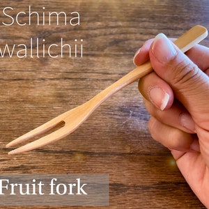 Puede incluir: Un tenedor de fruta de madera de color claro sostenido en una mano. El tenedor tiene dos púas y un mango plano. El texto "Schima wallichii" y "Fruit fork" son visibles en la imagen.