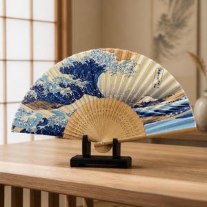 May include: A folding hand fan with a light wood frame and a printed design of a large blue ocean wave. The fan is displayed open on a black rectangular stand. The design includes white sea foam, a mountain, and vertical Japanese text characters.