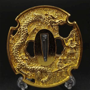 Puede incluir: Una tsuba circular de metal dorado con un diseño en relieve de un dragón largo y sinuoso y dos figuras con armadura tradicional. El centro contiene una abertura ovalada alargada y el borde exterior tiene cuatro muescas distintas.