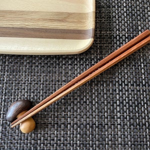 May include: Wooden chopsticks with a natural finish, resting on a small wooden chopstick rest. A wooden serving tray is in the background. The chopsticks are a reddish-brown color.