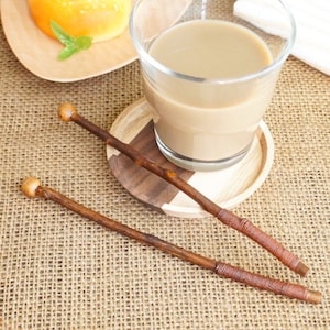 May include: Two handcrafted wooden wands with a light brown finish, each featuring a rounded knob at one end and wrapped detailing near the other. A glass of light brown liquid sits on a wooden coaster, with a pastry and drink in the background.