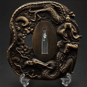 May include: A bronze-toned metal tsuba featuring an intricate relief design of a coiled dragon and a figure in traditional robes. The centre contains an oval opening with a tapered vertical slot, showcasing a dark, weathered metallic finish.