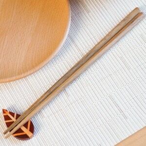 May include: Wooden chopsticks resting on a leaf-shaped chopstick rest. The chopsticks are light brown, and the rest is a combination of red and brown. A wooden plate is in the background, and the setting is on a white textured surface.