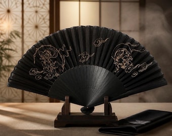May include: A black folding hand fan featuring gold-colored line art illustrations of two figures and stylized clouds. The fan is displayed open on a dark wooden stand, with a matching black fabric sleeve resting on the surface beside the stand.