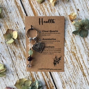 Op de afbeelding: Een bedelarmband met een zilveren hartbedel, een heldere kwartskristal, een groene aventurijnkristal en een paarse amethistkristal. De armband ligt op een bruine kaart met de tekst "Health" en beschrijvingen van de kristallen. De tekst bevat ook de website "HAPPYFAIRYgifts.etsy.com".