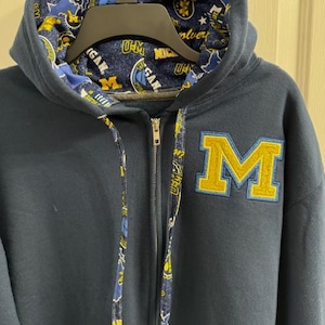 May include: Navy blue zip-up hoodie with a gold and blue "M" patch. The hood lining and drawstrings feature a patterned design with the words "Michigan" and "Wolverines". The hoodie has a full zipper and a hood.