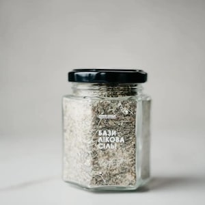 May include: A clear, hexagonal glass jar with a black lid, filled with a mixture of herbs and salt. The jar has a white label with text in a foreign language. The background is a soft, neutral gray.