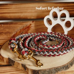 Könnte beinhalten: Eine geflochtene Hundeleine in Rot, Beige und Braun mit einem goldenen Metallverschluss. Die Leine ist auf einer Holzoberfläche mit einem weißen Pfotenabdruck-Ausschnitt dahinter aufgerollt. Der Text "Sofort lieferbar" ist im Hintergrund sichtbar.
