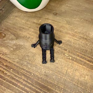 Peut inclure: Une cl&eacute; &agrave; douille noire imprim&eacute;e en 3D avec des bras et des jambes, ressemblant &agrave; un robot. La cl&eacute; est pos&eacute;e sur une surface en bois, avec un objet d&eacute;coratif blanc et vert en arri&egrave;re-plan.