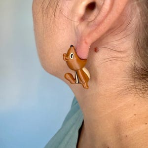 May include: Brown and white chipmunk earrings. The earrings are a cartoon-style design, with the chipmunk sitting upright. The chipmunk has a black nose and a white belly. The earrings are attached to a person's ear.