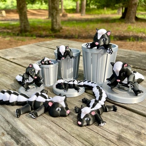May include: A set of black and white skunk figurines, some are in a small grey trash can, others are laying on a wooden surface.