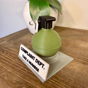 May include: A novelty desk accessory featuring a green grenade with a black top and a white sign that reads "COMPLAINT DEPT. TAKE A NUMBER."