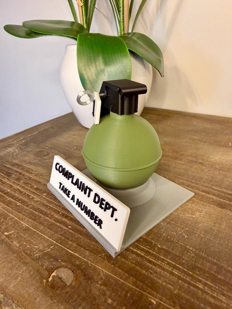 May include: A novelty desk accessory featuring a green grenade with a black top and a white sign that reads "COMPLAINT DEPT. TAKE A NUMBER."